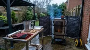 Winter BBQ gear setup in a UK garden featuring an insulated Masterbuilt smoker, windbreak, wireless meat thermometer, chimney starter, and heat-resistant gloves
